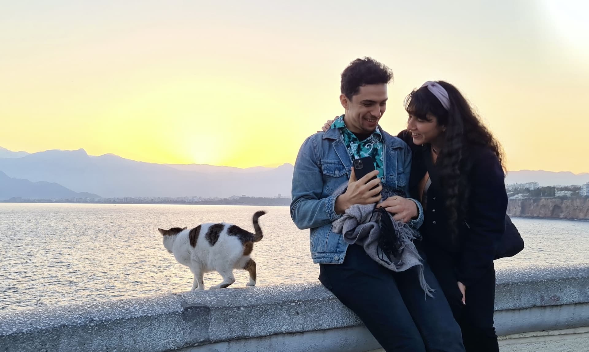 Me with my girlfriend and a cat sitting outside. Water and mountains are in the background.